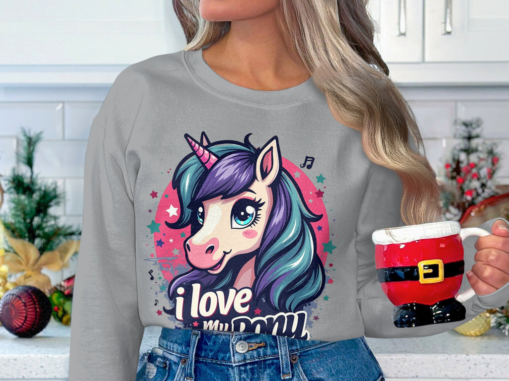 A person dons a Garment Graphics sweatshirt with a unicorn and I Love My Pony text, while holding a Santa-themed mug against a festively decorated white wall.