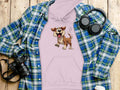 A Garment Graphics purple hoodie with a cartoon dog design sits atop a blue and green plaid shirt. Nearby are a camera, headphones, and a white coffee cup with a lid, all neatly arranged on a wooden surface.