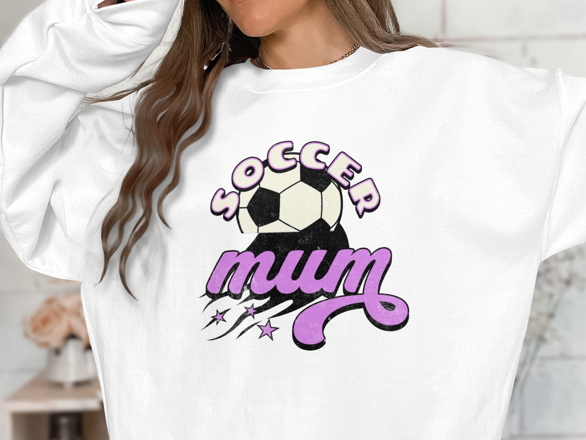A person wearing a Garment Graphics vintage-style white sweatshirt featuring Soccer Mum in bold, with soccer ball and purple design elements, their face is hidden by long, wavy brown hair.