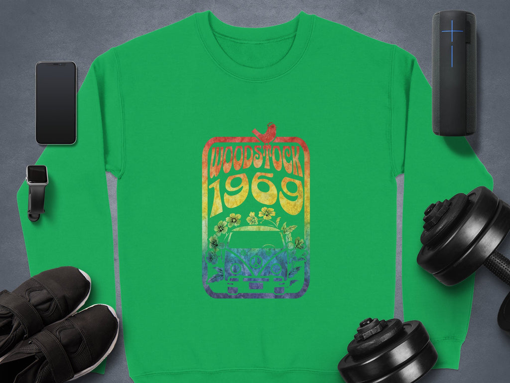 Garment Graphics green sweatshirt with a Woodstock 1969 rainbow design stands out, surrounded by retro-inspired items like a smartphone, smartwatch, portable speaker, sneakers, and dumbbells on the dark surface.