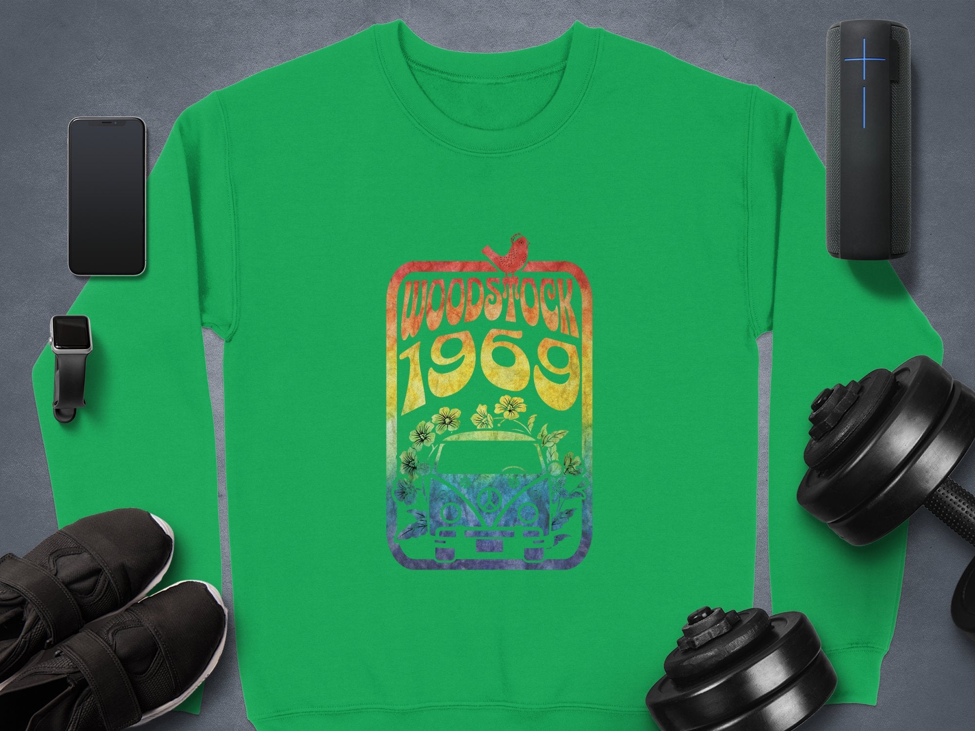 Garment Graphics green sweatshirt with a Woodstock 1969 rainbow design stands out, surrounded by retro-inspired items like a smartphone, smartwatch, portable speaker, sneakers, and dumbbells on the dark surface.