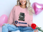 Someone sits cross-legged in a Garment Graphics pink graphic crewneck sweatshirt showcasing a Scania truck image and logo. Holding a red rose amidst heart-shaped balloons, their long, wavy hair cascades over ripped jeans—ideal for truck fans.