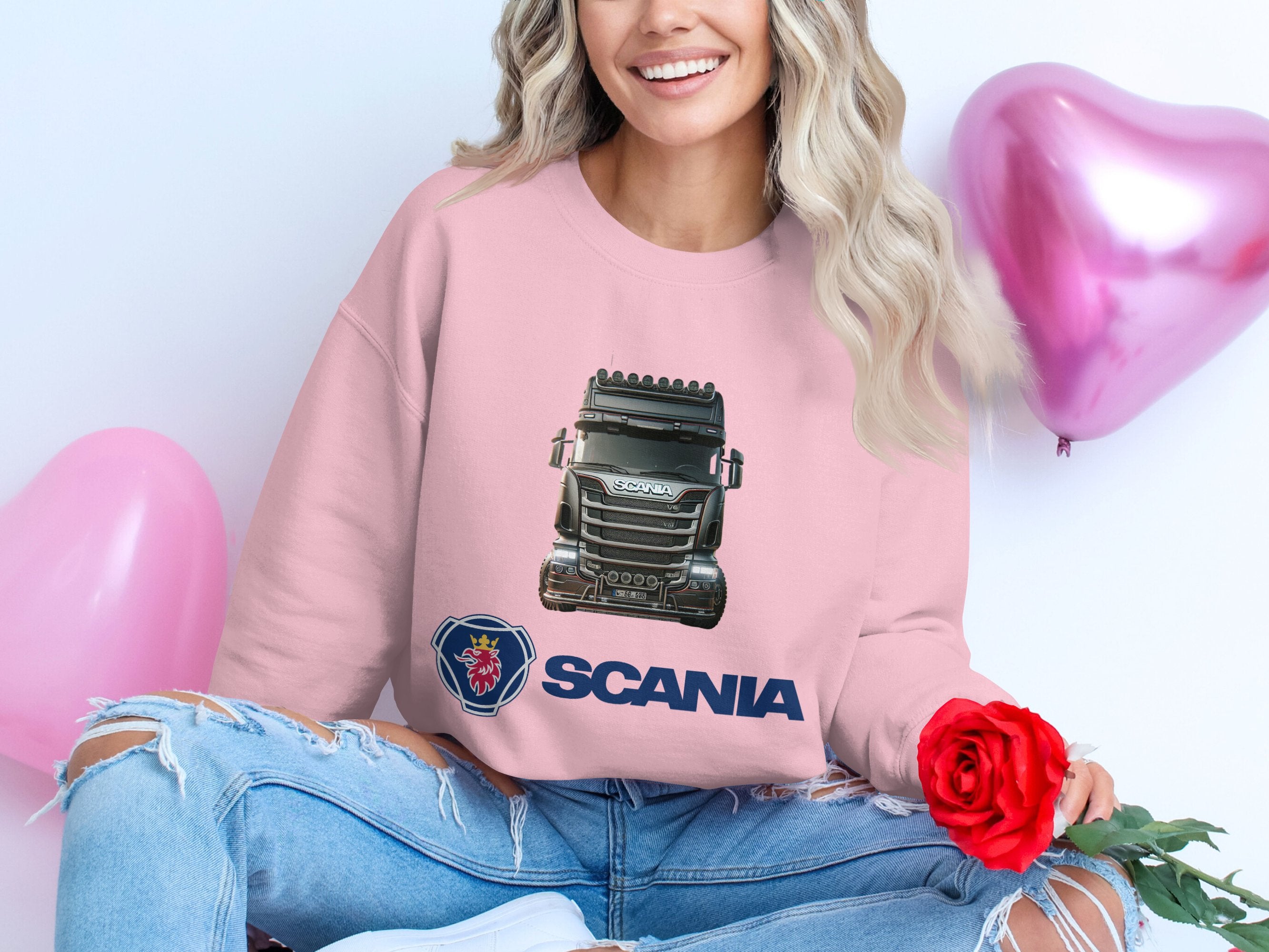 Someone sits cross-legged in a Garment Graphics pink graphic crewneck sweatshirt showcasing a Scania truck image and logo. Holding a red rose amidst heart-shaped balloons, their long, wavy hair cascades over ripped jeans—ideal for truck fans.