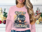 A person with long blonde hair stands indoors wearing a Garment Graphics sweatshirt featuring a whimsical truck and Keep On Trucking. Holiday decor surrounds them against white tiles and cabinets. This cozy unisex piece is perfect for everyone.