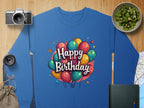 A blue Garment Graphics Happy Birthday unisex sweatshirt with colorful balloons is displayed on wood. Nearby are a camera, lens, sliced kiwi in a bowl, burlap bag, closed book, and a map—ideal for capturing festive moments.