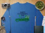 A Garment Graphics sweatshirt in blue says Happy St. Patricks Day with a green truck and shamrocks. Nearby, a camera, lens, bag, sliced kiwi, and book lie on wooden surfaces with part of a map visible.
