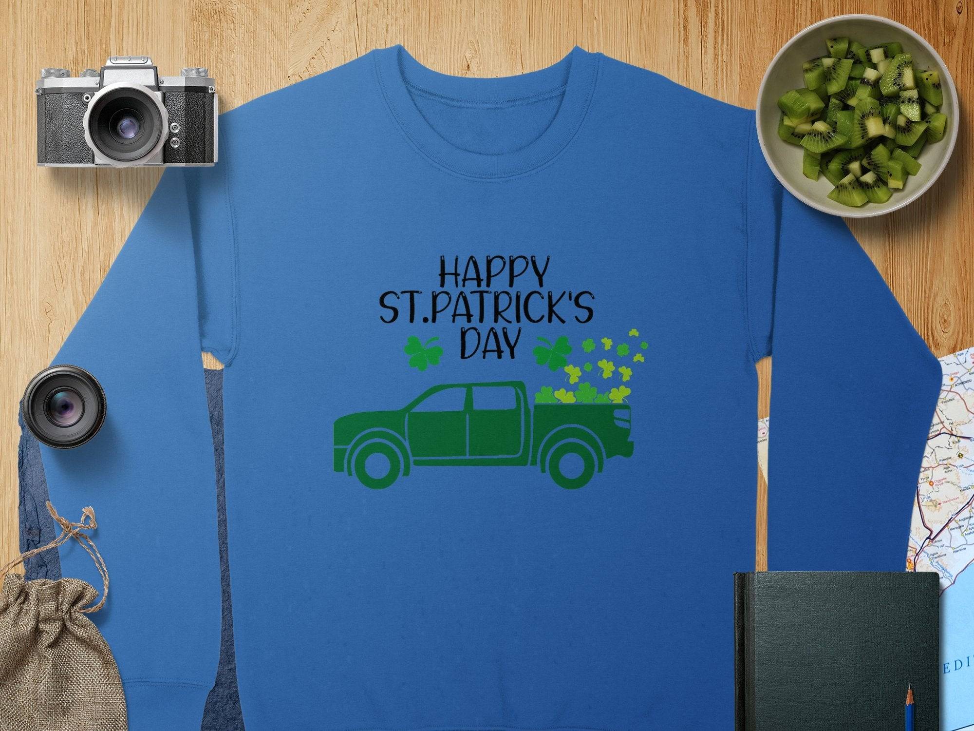 A Garment Graphics sweatshirt in blue says Happy St. Patricks Day with a green truck and shamrocks. Nearby, a camera, lens, bag, sliced kiwi, and book lie on wooden surfaces with part of a map visible.