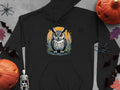 A black Garment Graphics hoodie with a fierce owl illustration on a circular background rests on a gray surface, surrounded by Halloween decorations like pumpkins, a skeleton, bats, and spiders. Its crafted from medium-heavy fabric for comfort and durability.