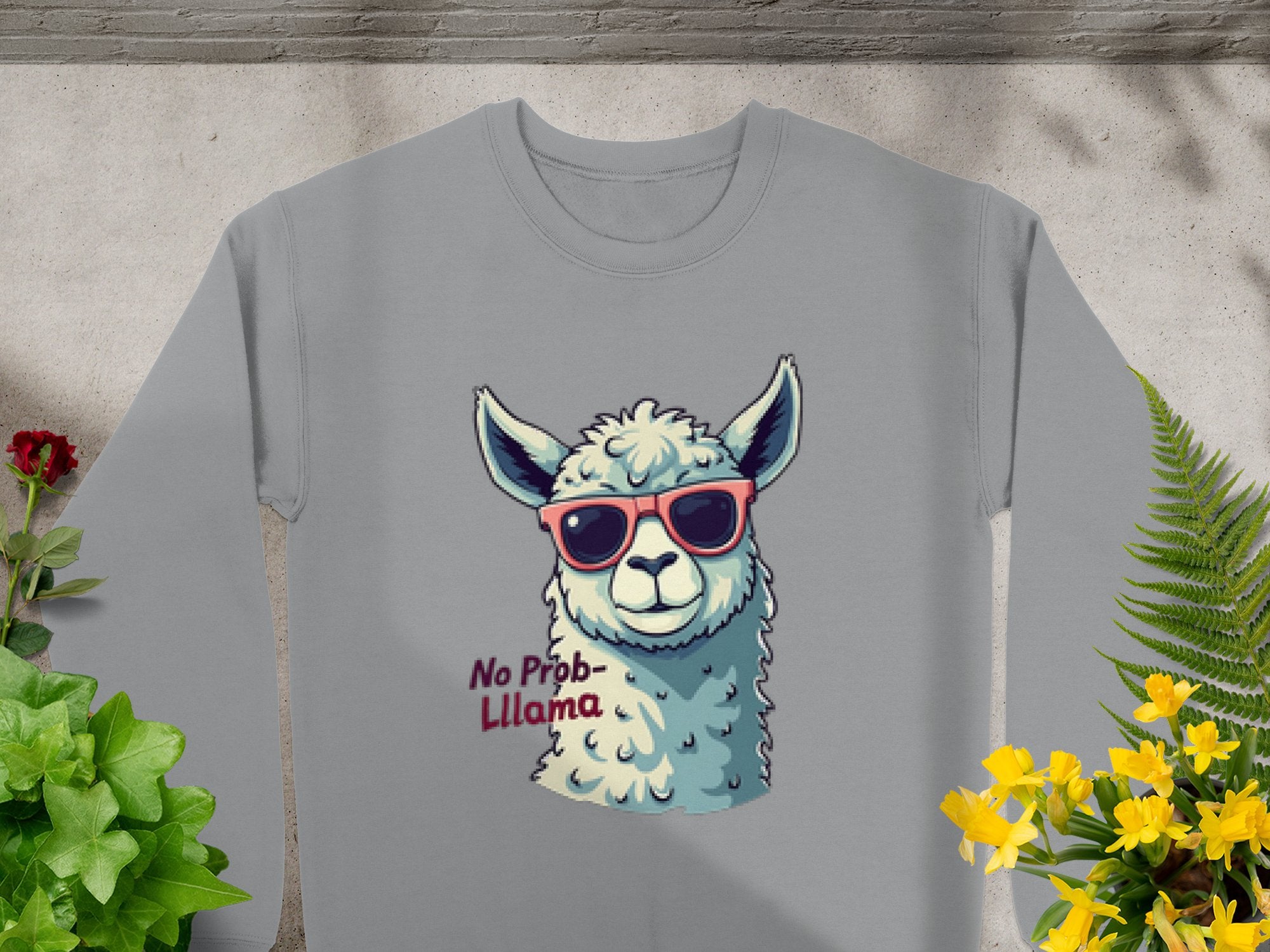 The Garment Graphics No Prob-Llama sweatshirt in gray features a playful llama with pink sunglasses and No Prob-llama text. It has a unisex fit and is styled against a concrete background with green and yellow plants.