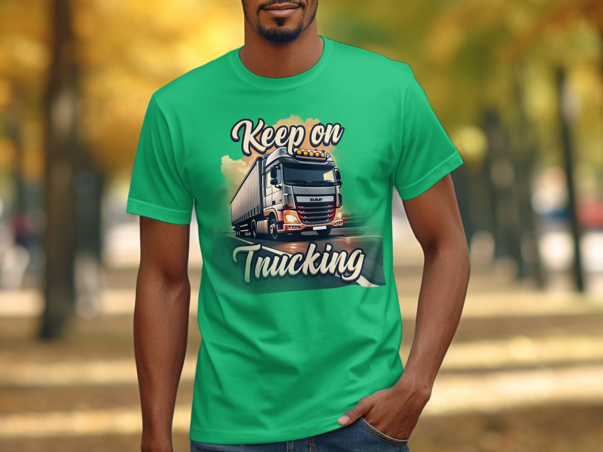 A person wears a green Garment Graphics t-shirt with a truck graphic and the phrase Keep on Trucking, ideal for truck enthusiasts. The background shows a blurred outdoor scene with trees and a path.
