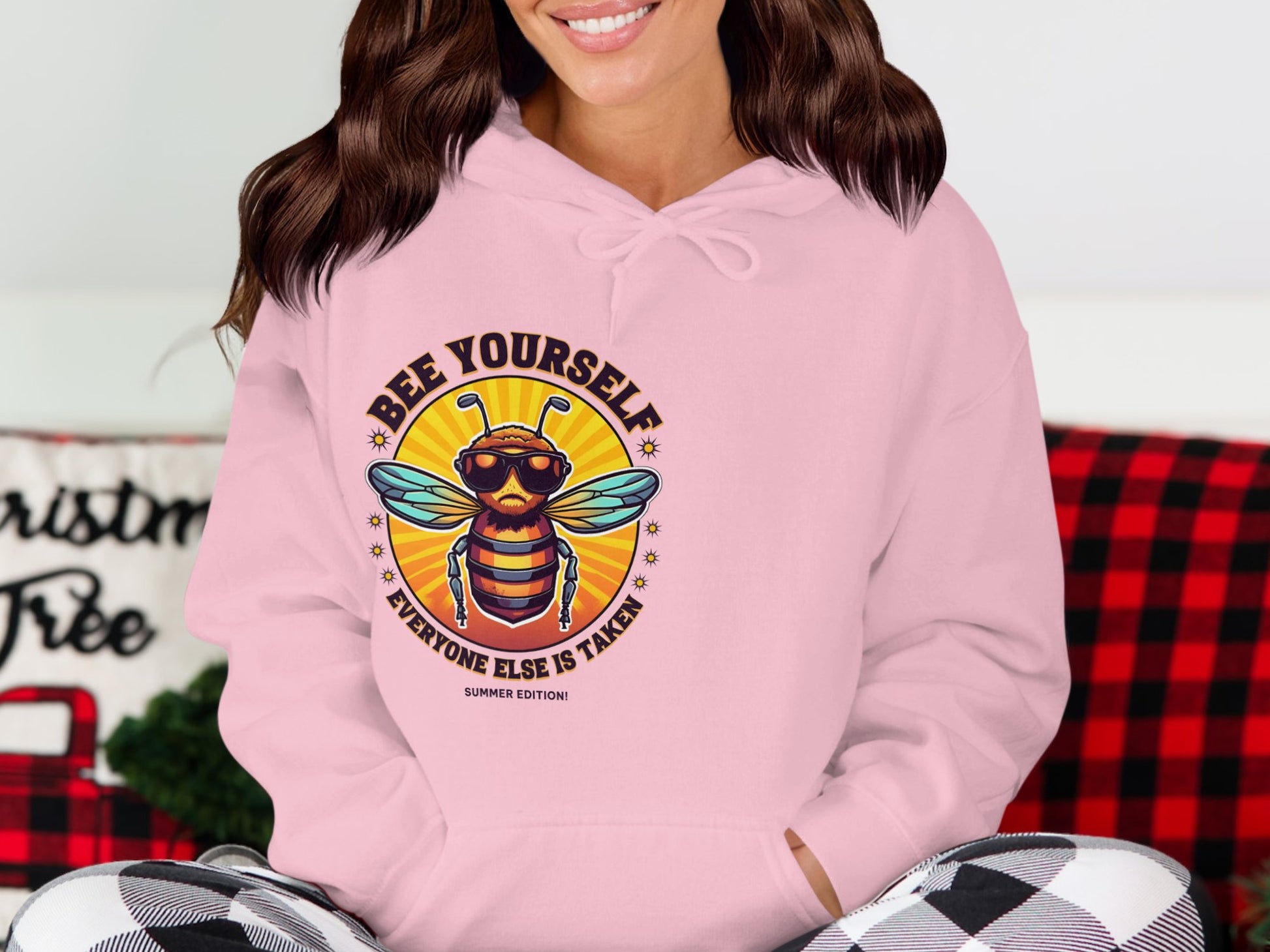 A person is in a Garment Graphics Summertime Edition Bee Yourself Hoodie, featuring a bee with sunglasses and the motto Bee Yourself, Everyone Else is Taken. Seated on a couch with a red and black plaid blanket, they effortlessly combine comfort and style.