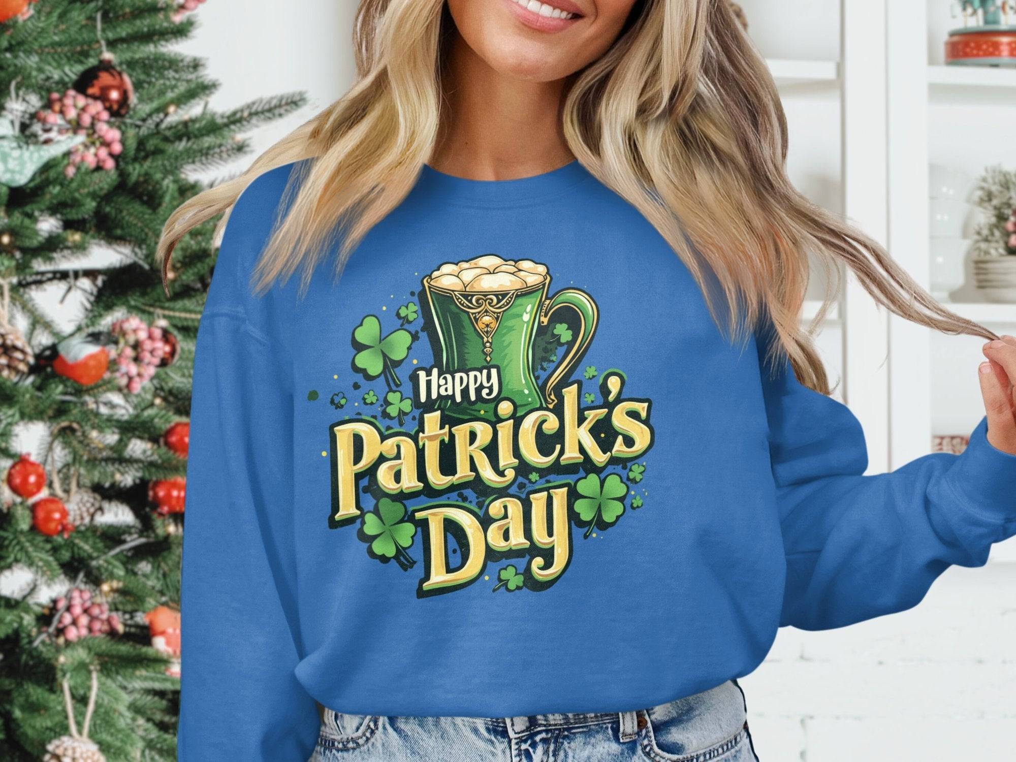 A person wearing a vibrant blue Garment Graphics sweatshirt featuring a Happy St. Patricks Day mug graphic stands before a decorated Christmas tree, surrounded by a white shelf adorned with plants and ornaments.