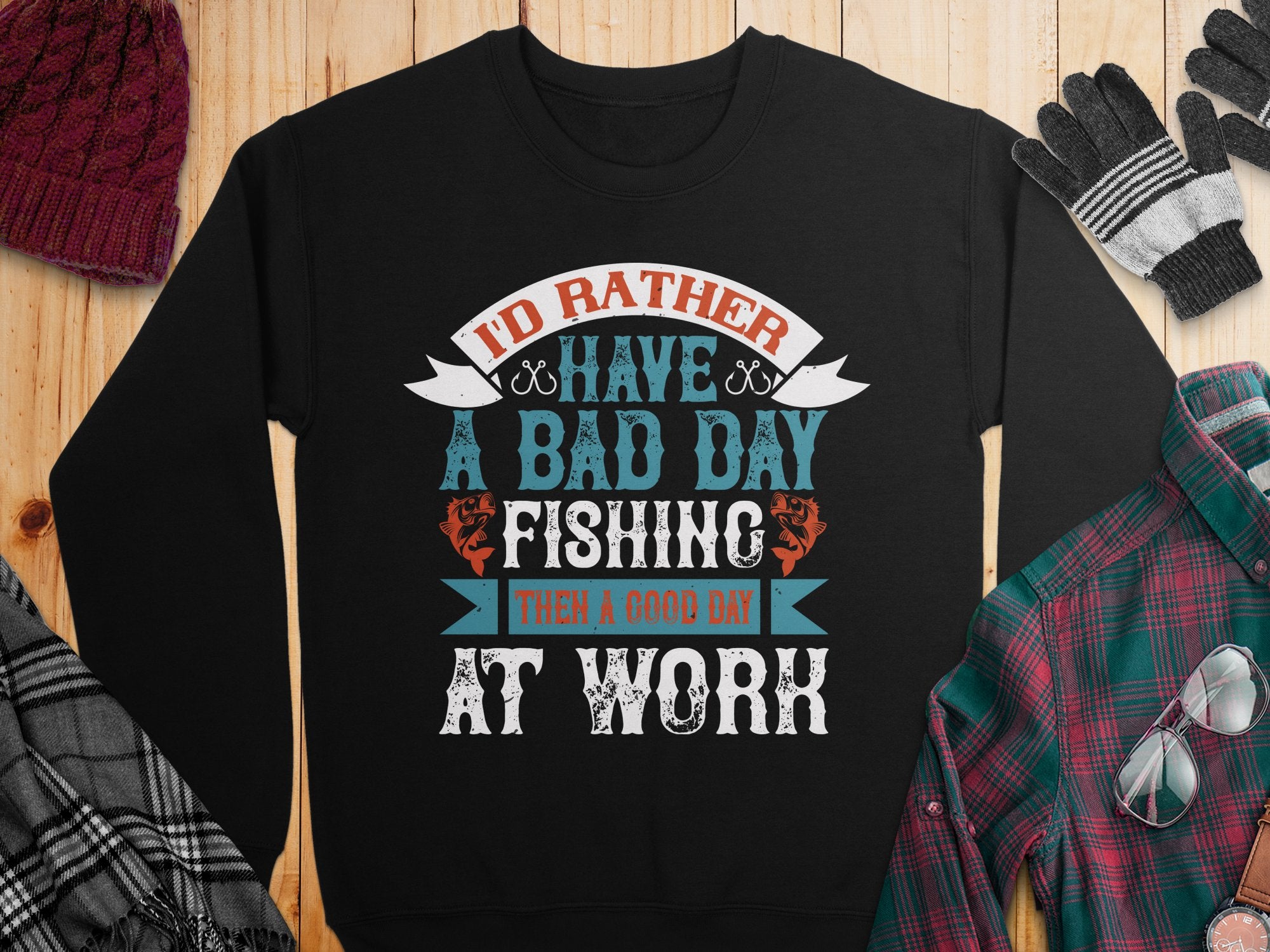 Garment Graphics presents a black unisex fishing sweatshirt with Id rather have a bad day fishing than a good day at work in mixed fonts and colors. Styled with a red beanie, plaid shirt, striped gloves, and glasses on wood—ideal for passionate fishers.
