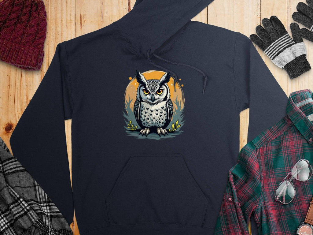 A Garment Graphics dark, medium-heavy hoodie with an illustrated owl rests on a wooden surface, surrounded by winter accessories: gray and black gloves, a red knitted beanie, a black and white plaid scarf, and a green and red flannel shirt.