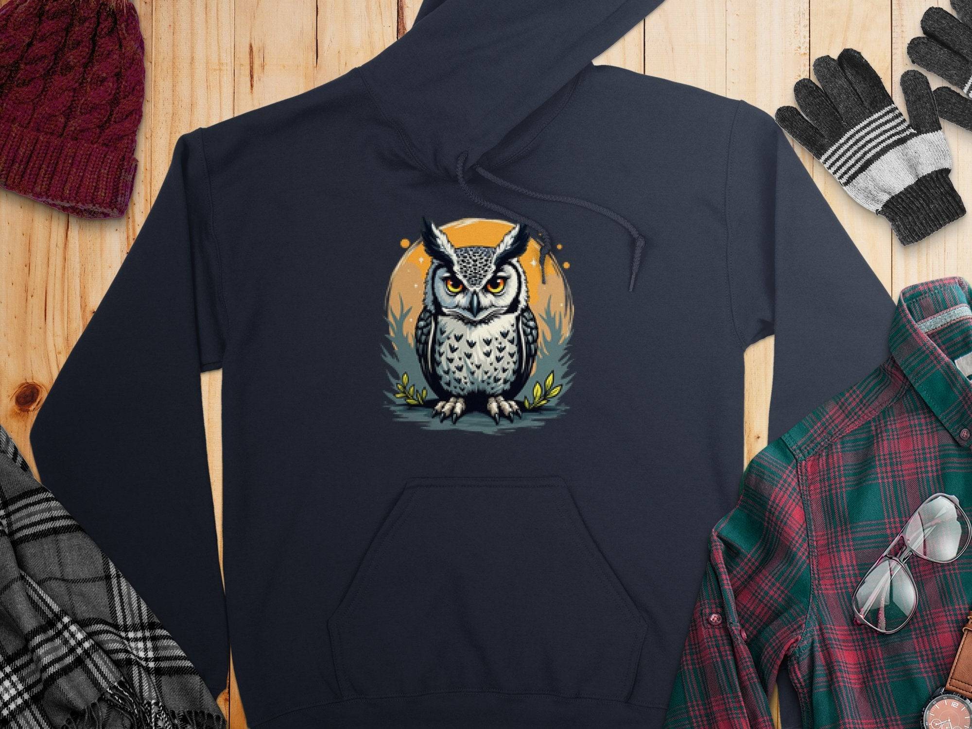 A Garment Graphics dark, medium-heavy hoodie with an illustrated owl rests on a wooden surface, surrounded by winter accessories: gray and black gloves, a red knitted beanie, a black and white plaid scarf, and a green and red flannel shirt.