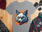 A 100% cotton gray t-shirt by Garment Graphics features a bold wolf print with bright orange eyes and an open mouth, accentuated by an orange paint splash. Styled on wood with a maroon beanie, gray gloves, plaid shirt, and checkered scarf for a complete look.