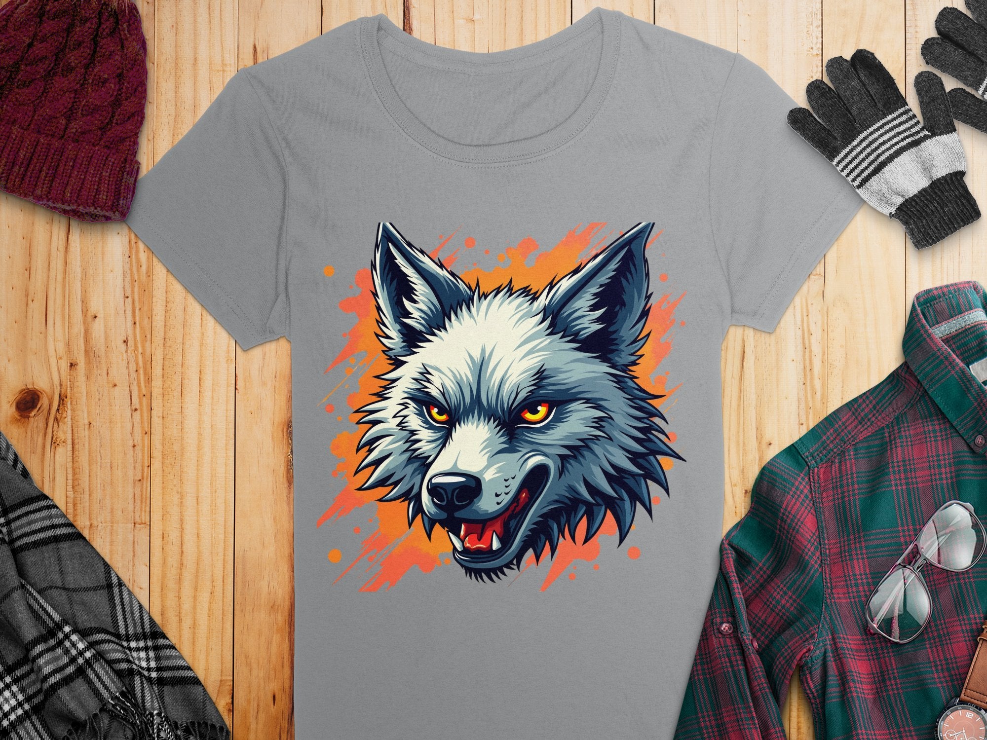 A 100% cotton gray t-shirt by Garment Graphics features a bold wolf print with bright orange eyes and an open mouth, accentuated by an orange paint splash. Styled on wood with a maroon beanie, gray gloves, plaid shirt, and checkered scarf for a complete look.