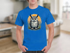Someone is wearing a Garment Graphics blue T-shirt showcasing a detailed owl graphic with large eyes, featuring an orange circle and stylized foliage in the background, set in a cozy, blurred interior space.