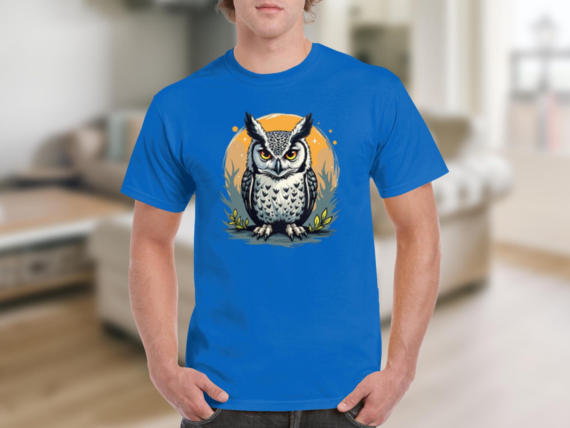 Someone is wearing a Garment Graphics blue T-shirt showcasing a detailed owl graphic with large eyes, featuring an orange circle and stylized foliage in the background, set in a cozy, blurred interior space.