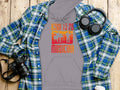 A flat lay on wood shows a Garment Graphics flannel shirt, gray Born To Be Musician unisex fashion hoodie with musician silhouettes, headphones, a camera, and a disposable coffee cup. Perfect for music lovers.