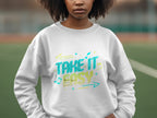 A person stands on a sports field, wearing a Garment Graphics unisex sweatshirt featuring Take It Easy in vibrant blue and yellow with arrows and music notes, making it pop against the blurred background.