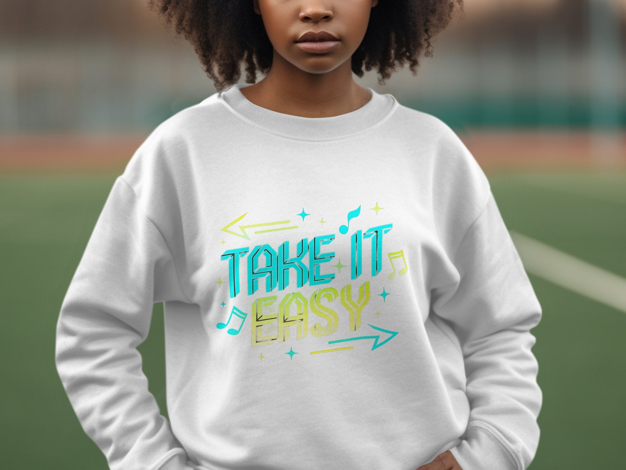 A person stands on a sports field, wearing a Garment Graphics unisex sweatshirt featuring Take It Easy in vibrant blue and yellow with arrows and music notes, making it pop against the blurred background.