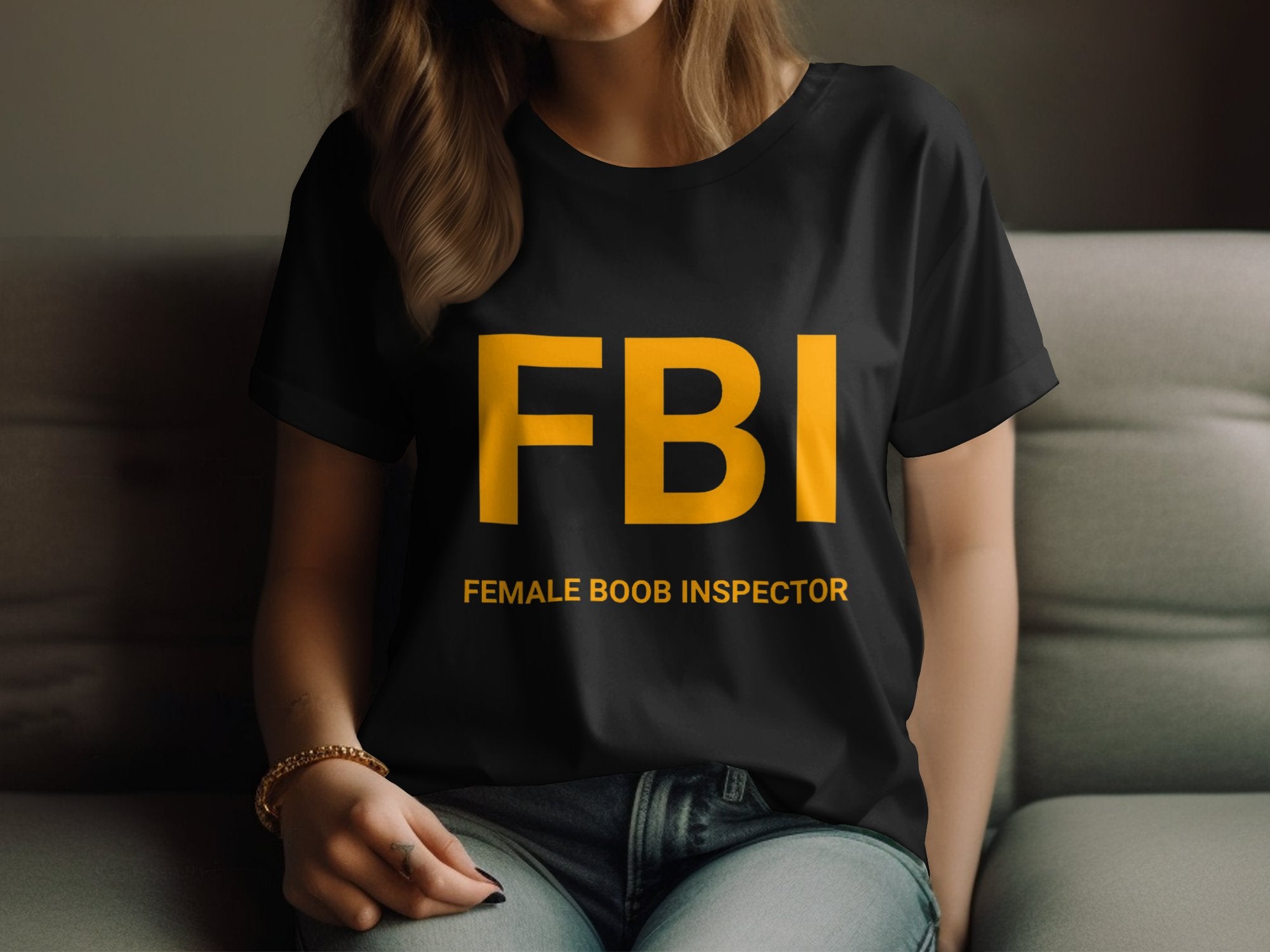 A person relaxes on a couch wearing a Garment Graphics FBI Female Boob Inspector T-shirt in womens sizing, featuring bold yellow letters for a gag gift vibe, while their other hand rests on their lap, highlighting their long hair.