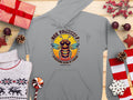 The Garment Graphics Bee Yourself Hoodie in gray showcases a bee with sunglasses and wings, encircled by the phrase Bee Yourself, Everyone Else is Taken, amidst gifts, a sweater, candy, pinecones, and holiday ornaments on a wooden surface.