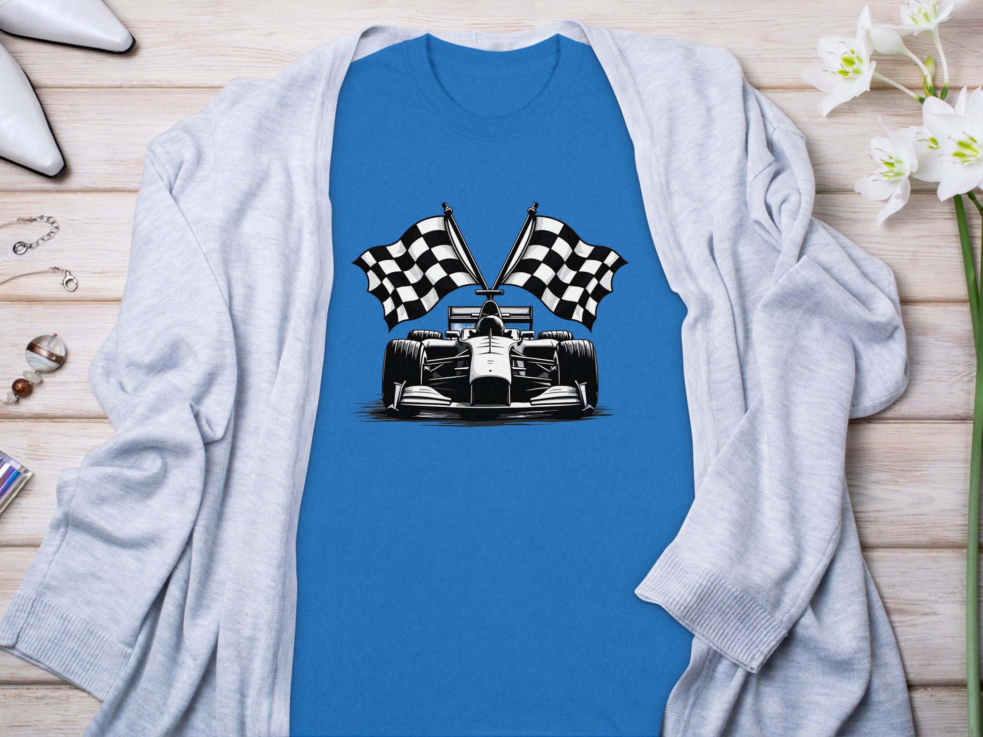Garment Graphics presents a blue graphic T-shirt with a black and white race car and crossed checkered flags design, embodying motorsport spirit. Styled chicly with a light gray cardigan, it is accentuated by white flowers and high-heeled shoes.