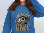 A woman wears a Garment Graphics blue graphic sweatshirt featuring an ornate eye design with intricate embellishments. Below the striking eye, GAZE is boldly printed. The unisex fit of the sweatshirt complements her long brown hair beautifully.