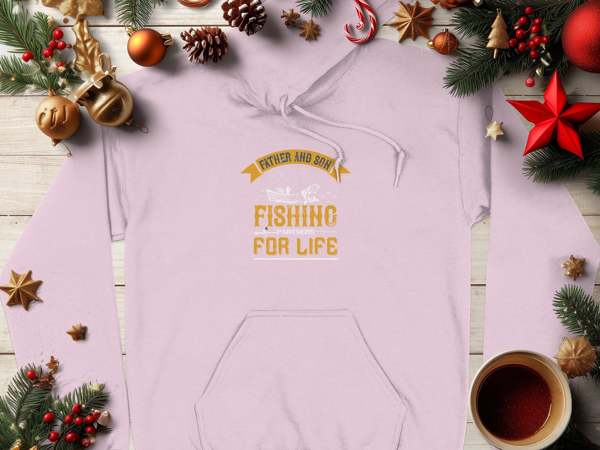 A Garment Graphics hoodie in light color with Father and Son Fishing Partners for Life is displayed on wood, surrounded by pine branches, cones, red ornaments, and a coffee cup. Its ideal for fishing fans amidst holiday decor during the festive season.