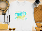 Flat lay featuring a casual white Garment Graphics sweatshirt with a Take It Easy graphic in blue and yellow, surrounded by a notebook, pen, plaid scarf, yellow quilted bag, sunglasses, jacket, and coffee cup on wood. Ideal for lovers of unisex comfort.
