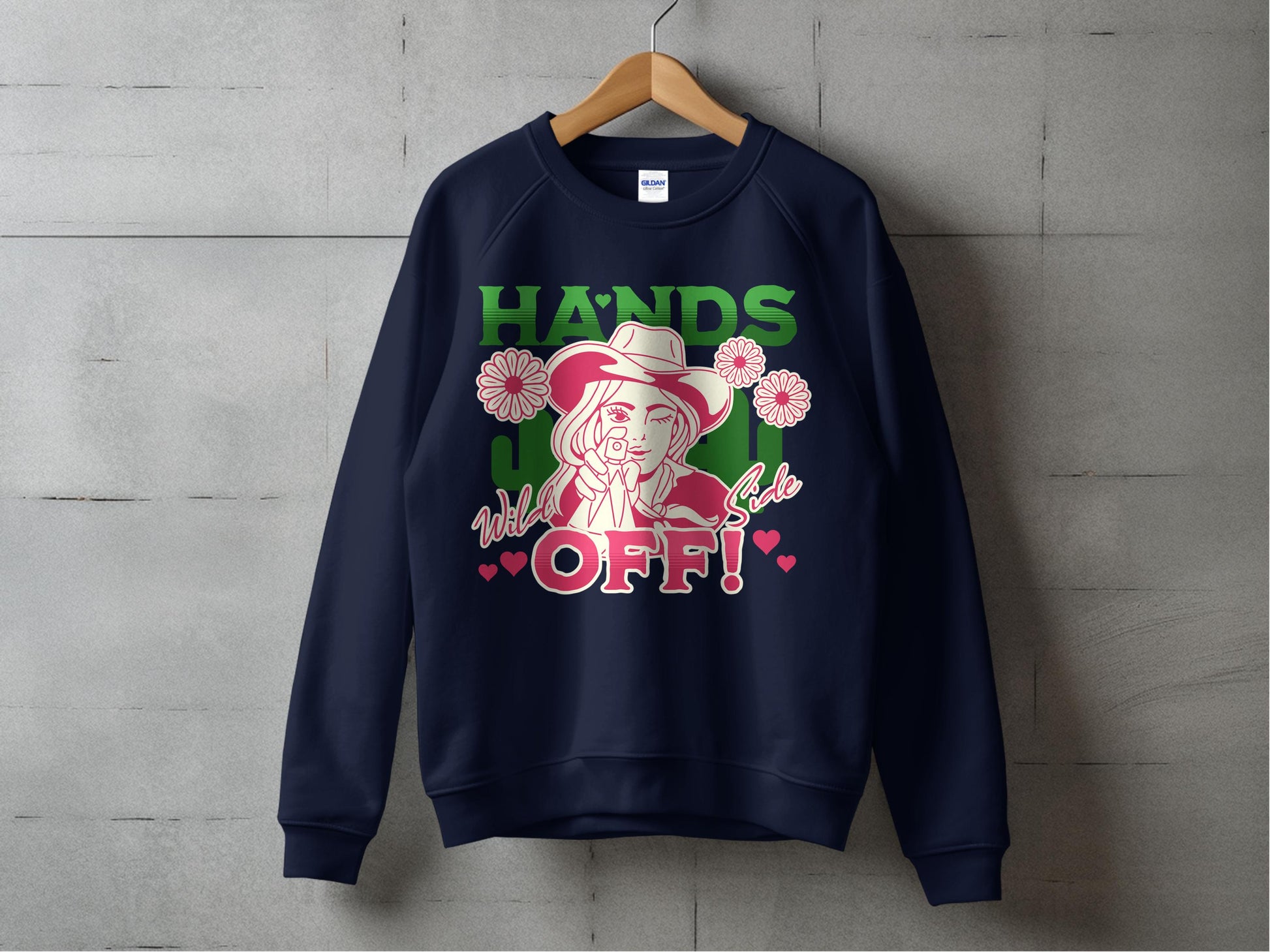 A navy sweatshirt from Garment Graphics Wild Side line hangs on a concrete wall, featuring vibrant HANDS OFF! graphics in green and pink with a cartoon cowgirl in a hat holding a lollipop, surrounded by pink roses and hearts.