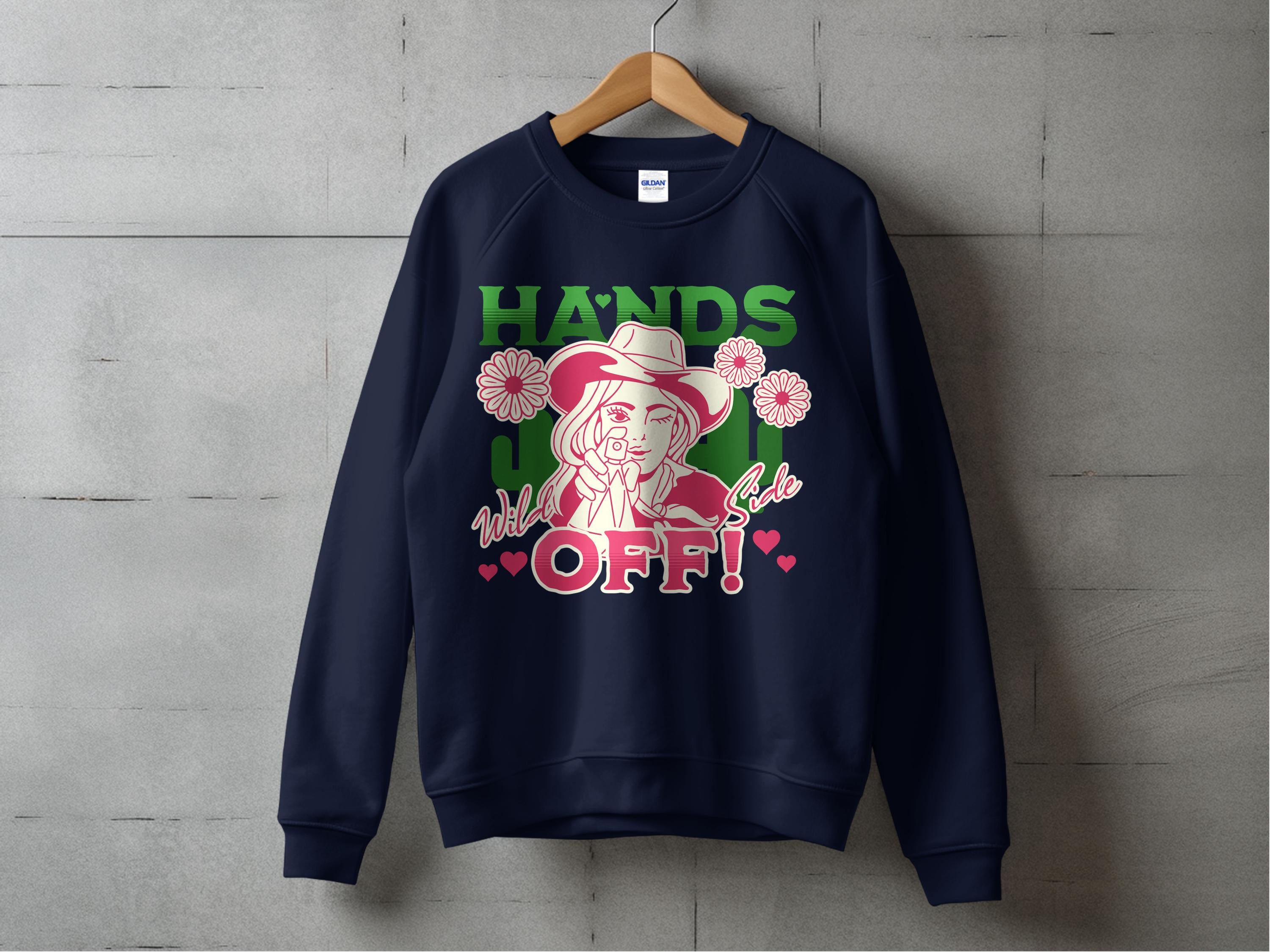 A navy sweatshirt from Garment Graphics Wild Side line hangs on a concrete wall, featuring vibrant HANDS OFF! graphics in green and pink with a cartoon cowgirl in a hat holding a lollipop, surrounded by pink roses and hearts.