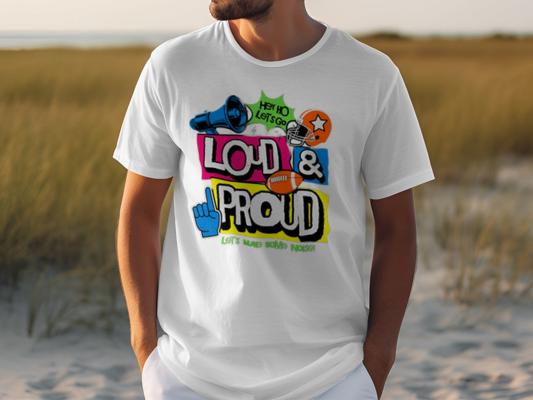 A person is wearing a light pink Garment Graphics T-shirt featuring a colorful Hey Ho Let’s Go Loud & Proud design in varied fonts. They stand on the beach, looking aside as the ocean and sandy shore provide an ideal backdrop, highlighting their passion.