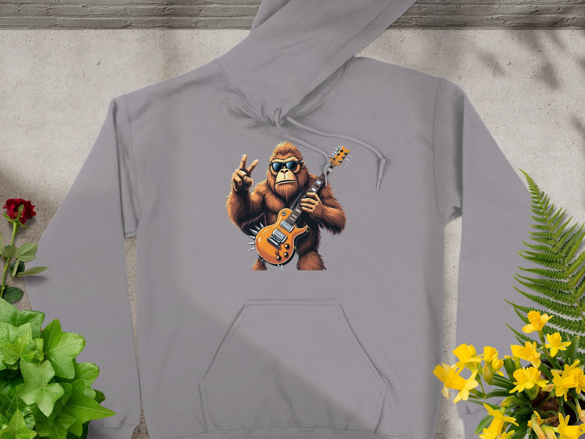 A Garment Graphics gray hoodie, featuring a sunglasses-wearing Bigfoot with a guitar and peace sign, is perfect for music and animal lovers alike. Styled among green foliage and a red flower, it offers both style and comfort.