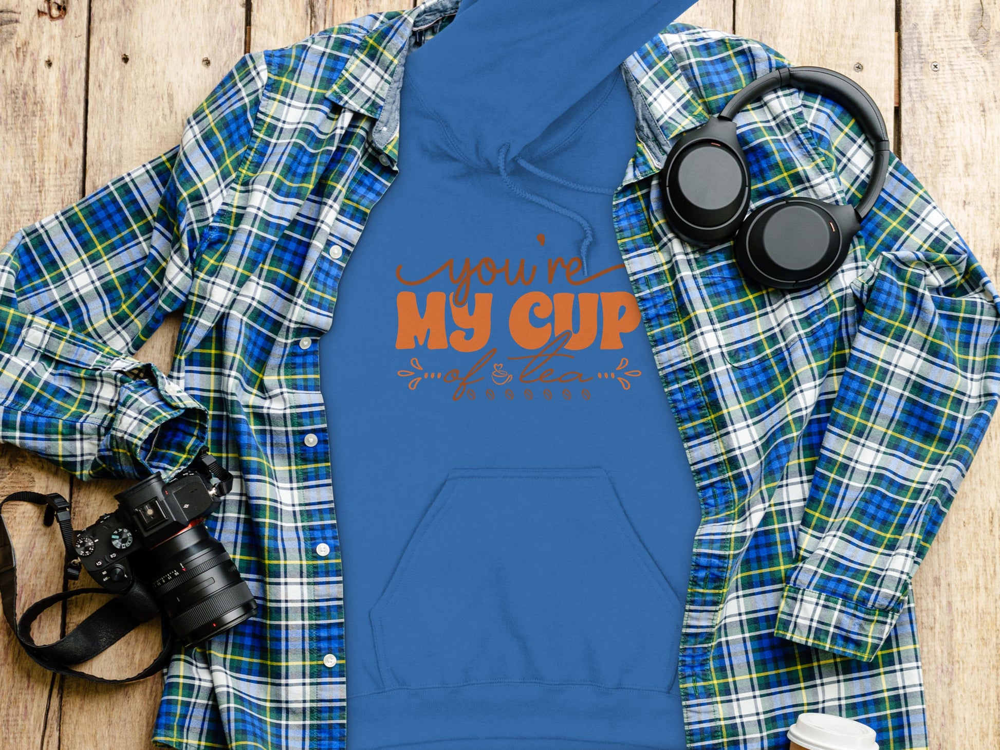 A blue Garment Graphics hoodie with the youre my cup of tea design in orange is placed on a wooden surface, accompanied by cozy moments with a blue and green plaid shirt, headphones, a camera, and a white paper cup.