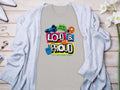 The Garment Graphics T-shirt features a vibrant Loud & Proud text with football graphics and phrases like Hey Ho Lets Go! Its ideal for family sizing and pairs well with a light gray cardigan, wristwatch, jewelry, and flowers.