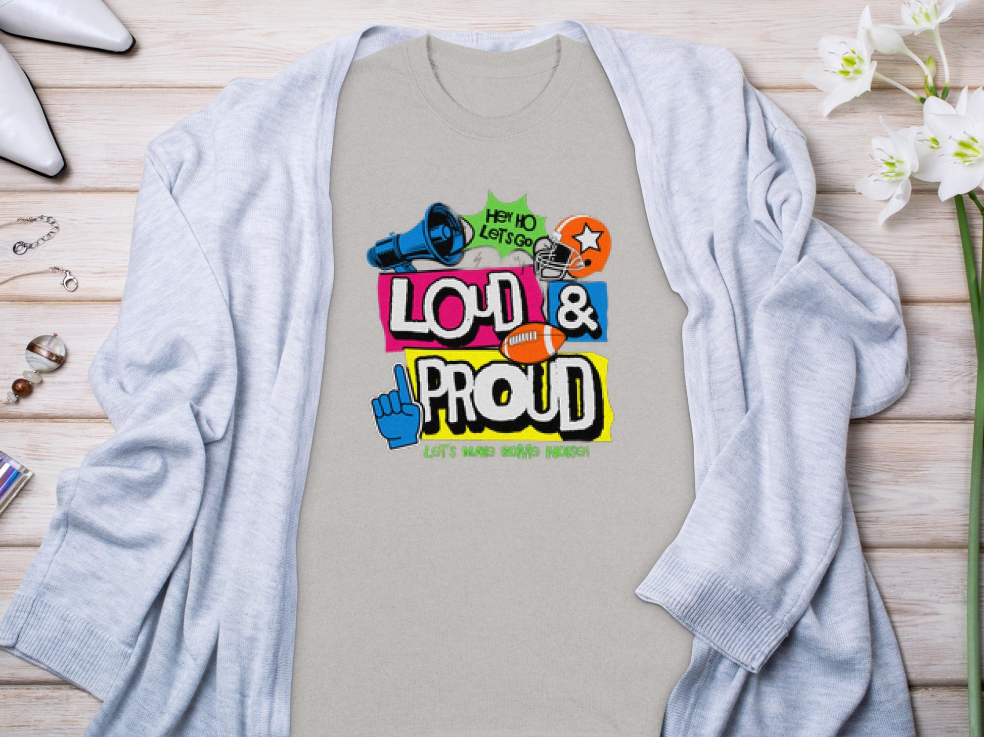 The Garment Graphics T-shirt features a vibrant Loud & Proud text with football graphics and phrases like Hey Ho Lets Go! Its ideal for family sizing and pairs well with a light gray cardigan, wristwatch, jewelry, and flowers.