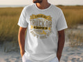 A person is wearing a white Garment Graphics T-shirt with gold and gray lettering that humorously states: If hes got golf clubs in his truck or a camper in his driveway, I dont hire him, standing outdoors amidst tall grass for a camping humor vibe.