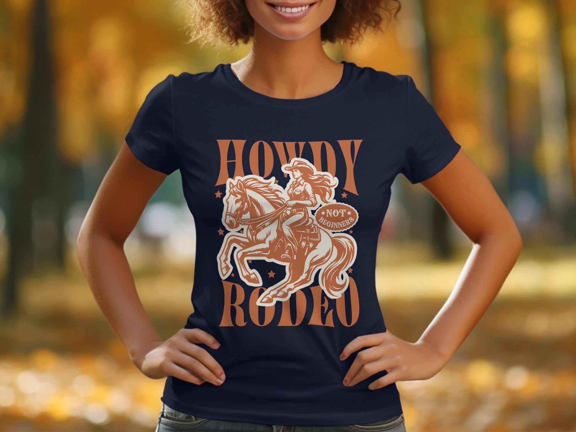 A woman in a navy blue Garment Graphics Rodeo T-shirt features an illustration of a horse rider with Howdy Rodeo and Not My First Rodeo text, set against a blurred outdoor scene with autumn leaves.