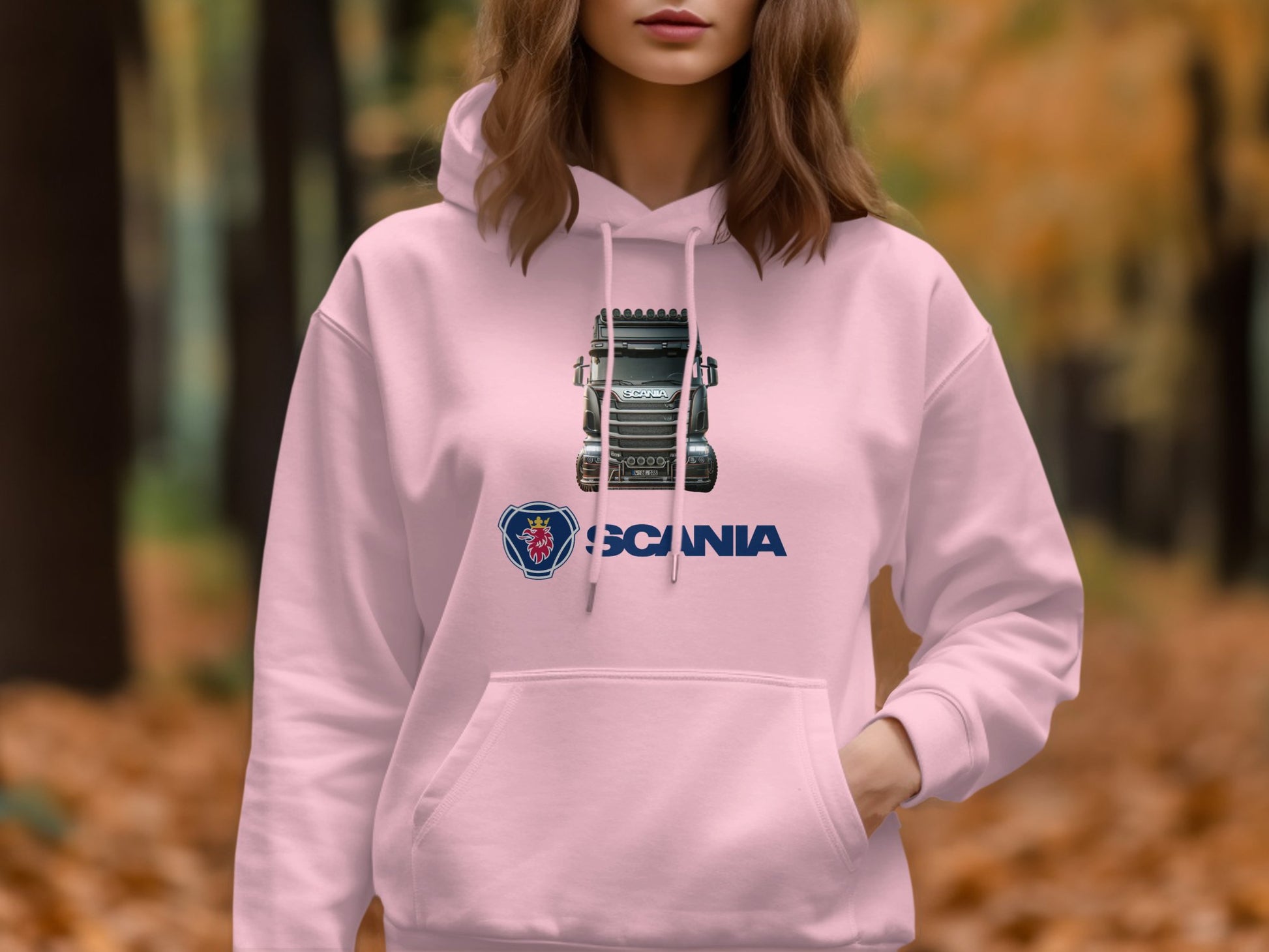 A truck enthusiast in a pink Garment Graphics hoodie flaunts a bold design with a truck and lion emblem, set against the perfect autumn forest backdrop with fallen leaves.