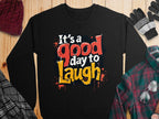 A black Garment Graphics sweatshirt with Its a good day to laugh in bold white, red, orange, and yellow fonts rests on wood. Nearby are a maroon knit hat, gloves, a plaid scarf and shirt, plus glasses.