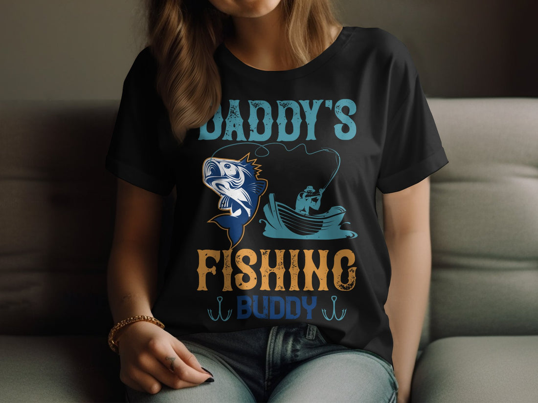 A Garment Graphics womens fishing shirt reads Daddys Fishing Buddy with a boat and fish illustration. Its displayed on a wooden surface with holiday decorations like wrapped gifts, pine cones, and ornaments.