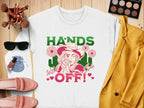 The Garment Graphics Hands Off Wild Side Sweatshirt is a white, loose-fit top featuring bold Hands Off! text and a woman in a hat surrounded by flowers. Styled with a yellow jacket, pink shoes, sunglasses, makeup brushes, and an eyeshadow palette on a wooden surface.