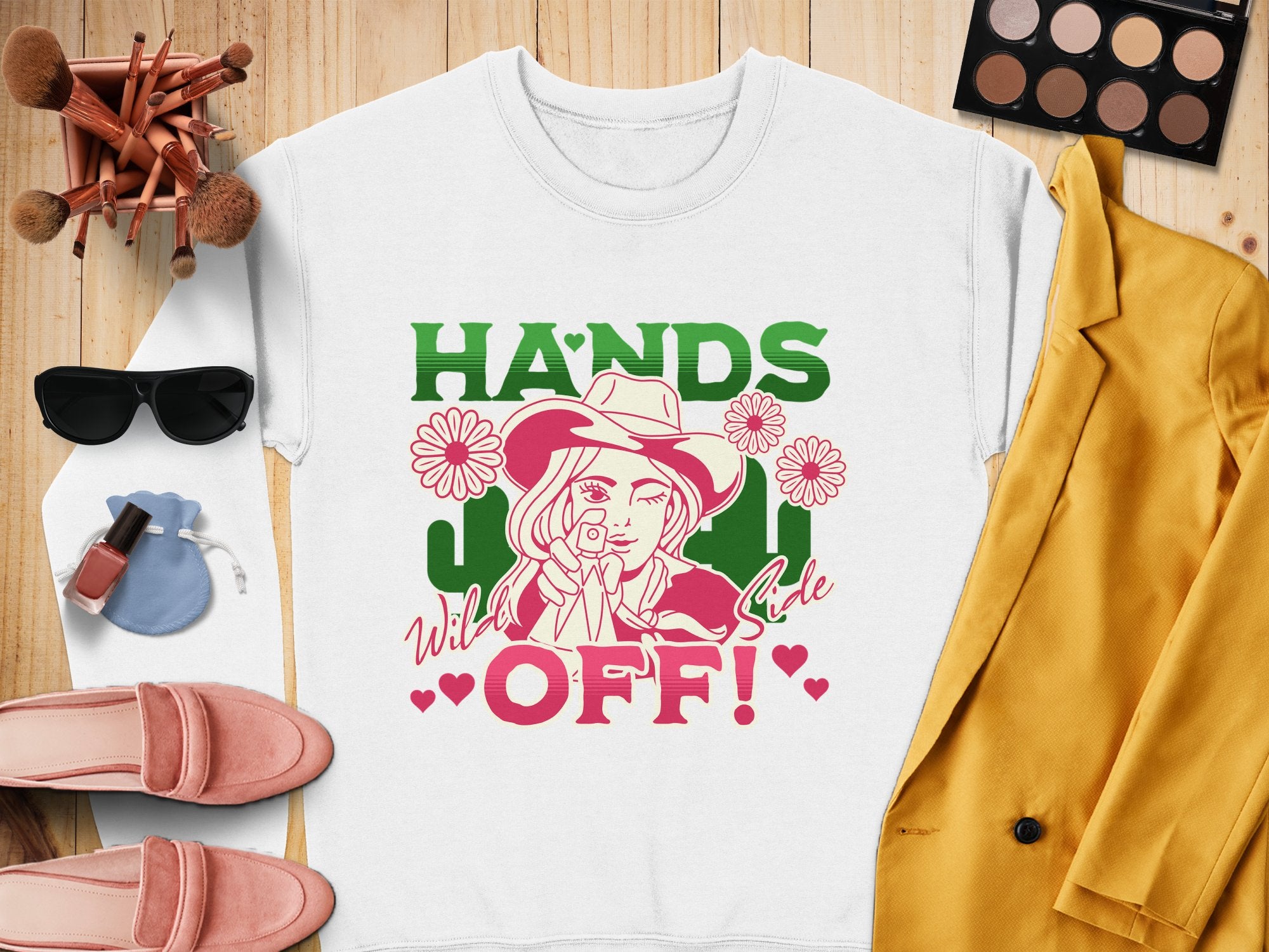 The Garment Graphics Hands Off Wild Side Sweatshirt is a white, loose-fit top featuring bold Hands Off! text and a woman in a hat surrounded by flowers. Styled with a yellow jacket, pink shoes, sunglasses, makeup brushes, and an eyeshadow palette on a wooden surface.