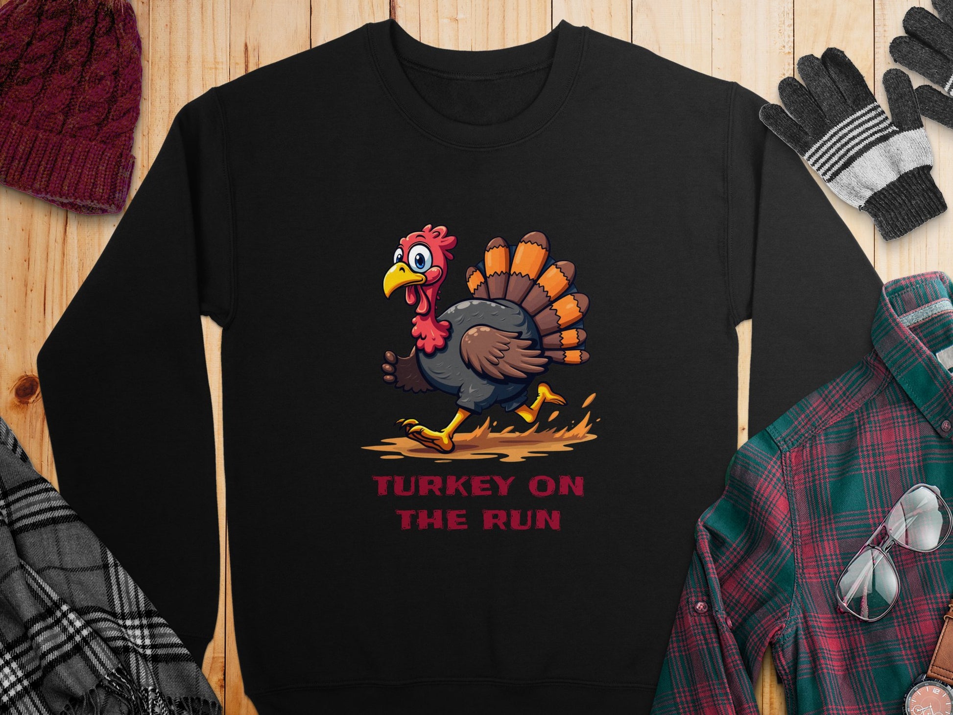 Garment Graphics offers a unisex black sweatshirt featuring a cartoon turkey running with the caption Turkey On The Run in bold red text. Styled with a red beanie, plaid shirt, black gloves, and glasses on rustic wood for a complete humorous look.