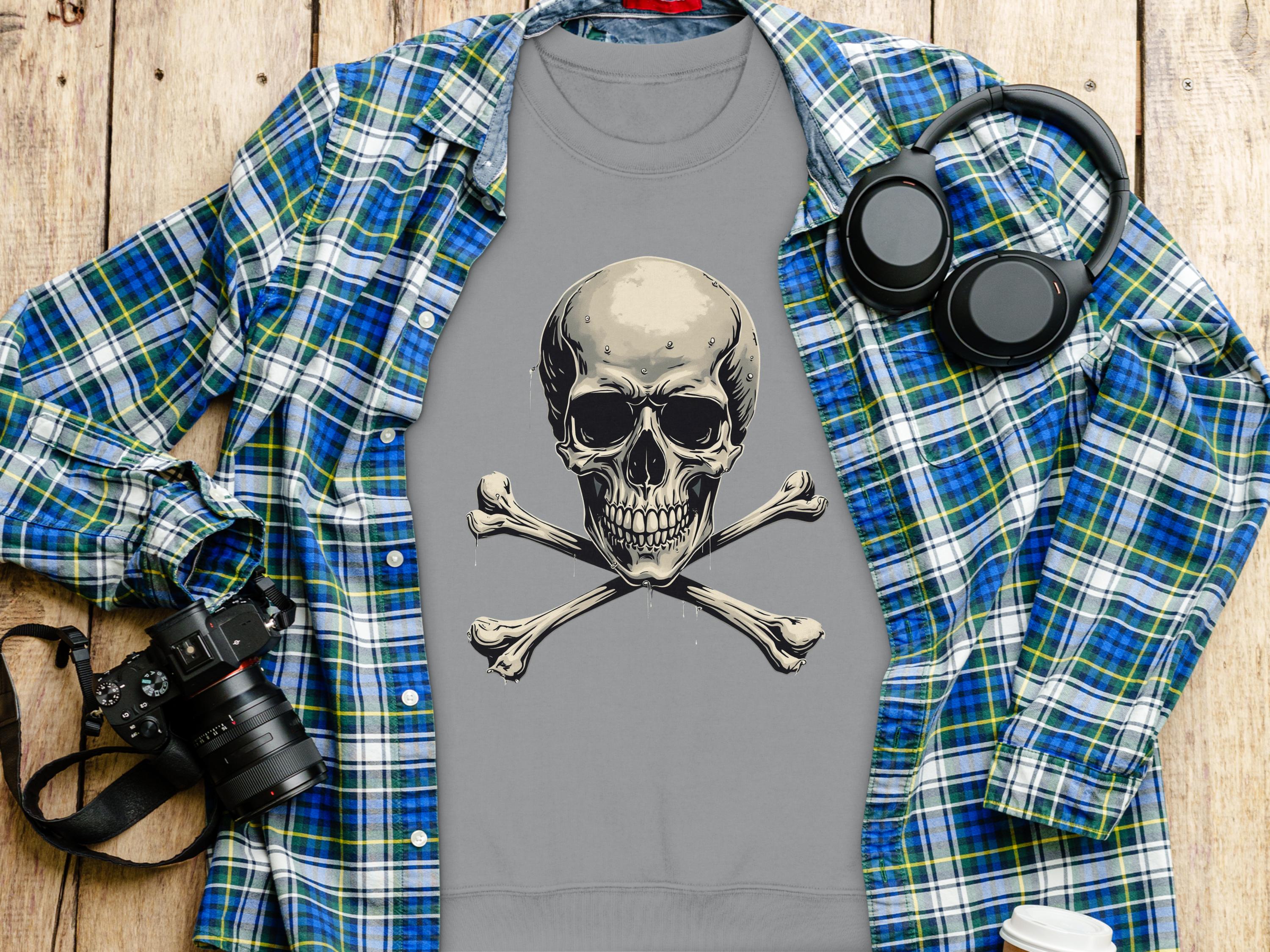 A Garment Graphics gray t-shirt featuring a bold skull and crossbones design tops a blue-green plaid shirt. Nearby are a black camera, sleek black headphones, and a white insulated drink container, all set against a wooden background.