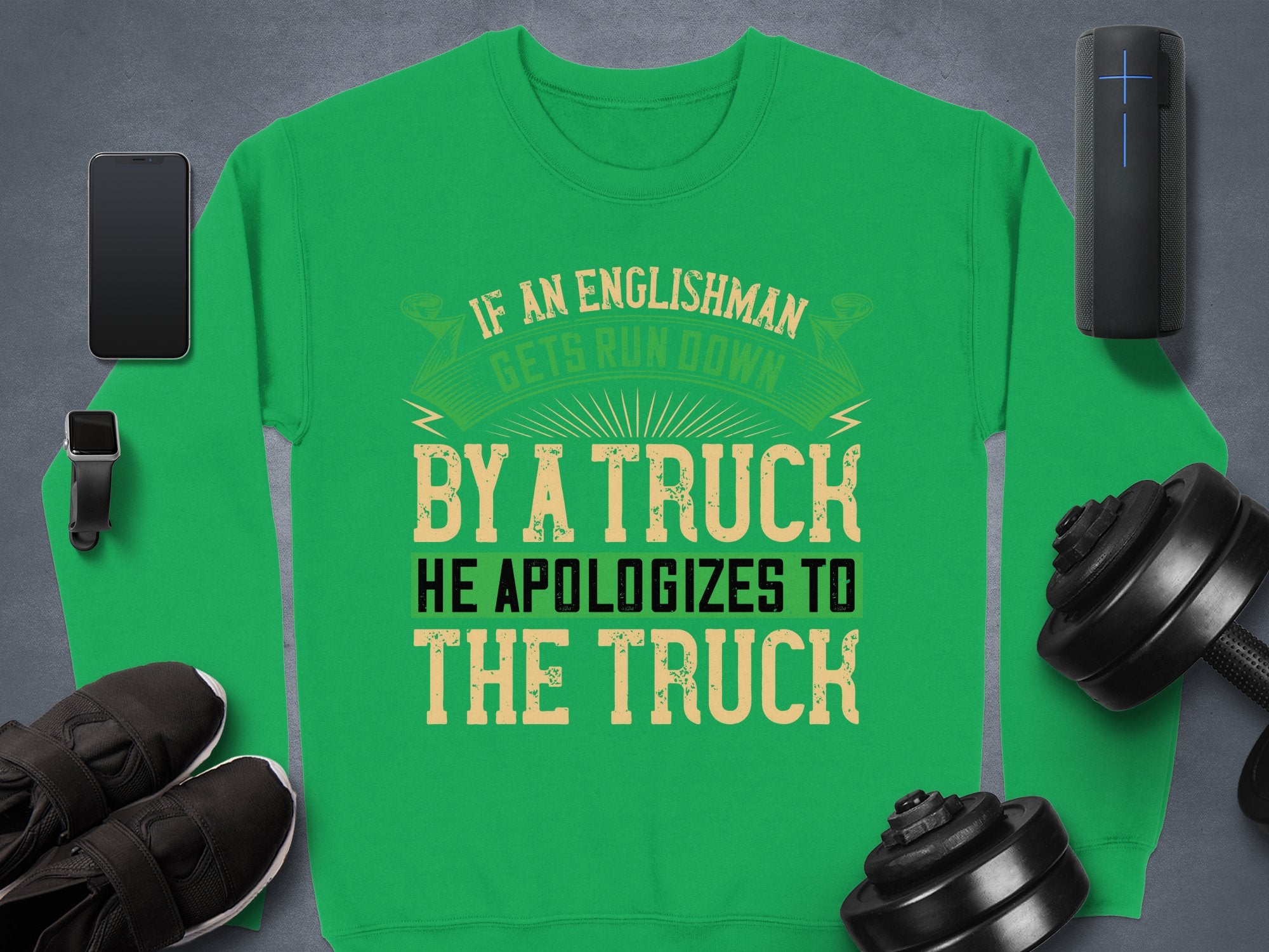 Garment Graphics offers a unisex green sweatshirt with a bold, humorous design: If an Englishman gets run down by a truck, he apologizes to the truck. Its displayed on a gray surface surrounded by a smartphone, smartwatch, portable speaker, shoes, and dumbbells.