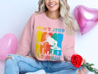 A person with long blonde hair beams joyfully, wearing a vibrant DJ-themed pink sweatshirt with the slogan Dont Stop the Music by Garment Graphics, perfect for music lovers. They sit cross-legged holding a red rose, surrounded by pink heart-shaped balloons.